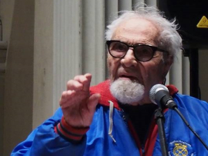 Herman Benson at podium in blue jacket with arm raised