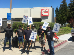 Members of Machinists Local 1546 stand in front of their workplace holding picket signs.