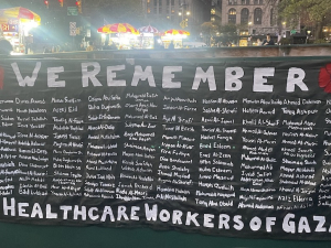 A black banner with white letters lists 160 health care workers killed in Gaza. It is titled “We Remember”.