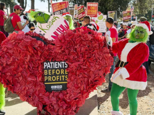 Two people in green Grinch suits hold a giant paper-mache heart that contains a "patients before profits" sign. Behind them an NUHW Kaiser picket line crowd is visible.