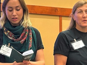 Two women in dark t-shirts stand side by side, one reading from a thick booklet