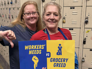 Two white women stand together in front of a wall of lockers, presumably the non-public area of a grocery store, holding a printed blue and yellow sign that reads "Workers' needs vs. grocery greed," with symbols of a fist and Mr. Moneybags. The woman on the left is giving the "thumbs-down" sign. Both smile calmly.