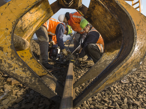 Workers in orange vests and hardhats crouch around a track. They are framed by a big yellow piece of equipment in the foreground. The day is very sunny.