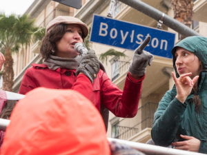 A woman speaks at a strike rally while another woman interprets in sign language. Behind them is a palm tree, an apartment buidling or hotel, and a street sign "Boylston"