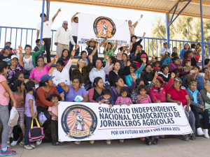 A group of Mexican women farmworkers sit in several rows behind the SINDJA banner.