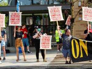 Eight picketers, who appear mostly young and white, stand in front of New Seasons. Their screen-printed picket signs say "New Seasons Labor Union, On Strike, Unfair Employer, Do Not Patronize" in red ink with flourishes. Two hold a black banner with yellow letters, only partly visible but appears to say "On strike." They all stand near the curb, facing traffic, and one has a bullhorn.