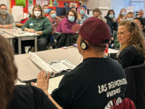 People sit in a classroom. Most are looking attentively at a person in the foreground who must be speaking, but has his back to us. He is wearing an interpretation headset and the back of his T-shirt says "Los Deliveristas Unidos," Spanish for united delivery workers.