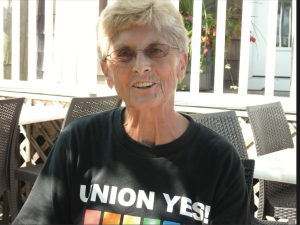 A smiling woman is pictured with short blond hair and a black Union Yes t-shirt with a rainbow on it.