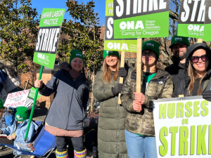 Women stand on a sunny day, wearing big jackets, holding green printed ONA picket signs that say "Unfair Labor Practice Strike" and "Nurses on Strike." A handmade sign visible in the background says "Burnout caused this turnout."