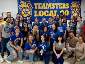 A group of 25 blue-shirted nurses pose happily in front of a Teamsters Local 90 banner