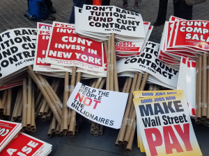 stack of picket signs including "don't raise tuition, fund CUNY contract," "good jobs now, make Wall Street pay," "CUNY invest in New York," "help the people, not the billionaires," and "CUNY needs competitive salaries"