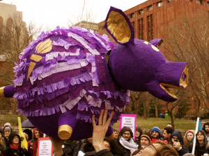 A crowd surrounds a large festive purple piggy-bank piñata suspended above them. It is labeled NYU and is in school colors, purple and gold.