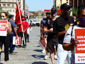 Masked people stand outside holding red printed signs. Visible sign in foreground says: "Solidarity with Sanitation Workers! Hazard Pay and PPE Now!"