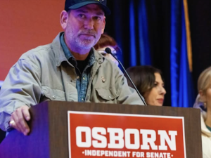 A man in a ball cap stands behind a lectern that says Osborn for Senate