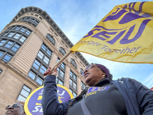From a heroic low angle, we look up at a Black woman marching in a purple SEIU BJ shirt. The yellow SEIU flag she carries is billowing in the wind, mirroring the curve of the building behind her, against the blue sky. We can see the corners of other picket signs--she is part of a dense crowd.