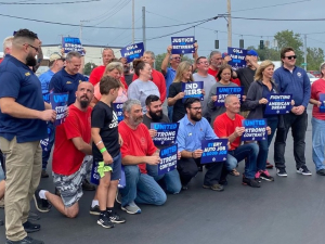 A group of workers in red, blue, or grey shirts hold signs saying “United for a Strong Contract,” and “COLA and Fair Pay Now”  