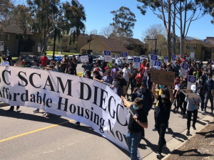 A group marches down the street behind a banner reading "UC Scam Diego: Affordable Housing Now."