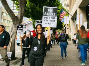 A woman holds a bullhorn and a picket sign that says "On Strike for a Fair Contract," surrounded by dozens of picketers holding similar signs and marching in a circle on the sidewalk.