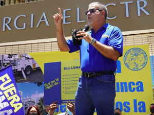 Man in blue shirt with right arm raised and left arm holding microphone addressing crowd, Fuera LUMA flag visible in foreground, building of Puerto Rico Electric Power Authority (PREPA)