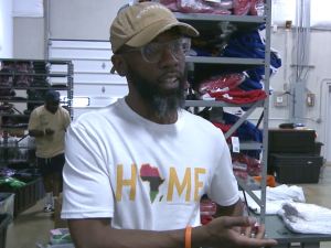 A Black man in a baseball cap and a T-shirt that says "Home" with the shape of Africa for the "O" speaks to the camera. He is standing in a warehouse, in front of shelves stacked high with folded apparel . Another Black man can be seen behind him, working.