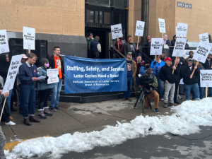 Postal workers rally outside a post office. There is some snow on the ground. A big blue banner says "Staffing, Safety, and Service: Letter Carriers Need a Raise! NALC Branch 9, Minneapolis." Printed picket signs say "End Mandatory Overtime," "First Class Service, First Class Pay," and "Fair Contract Now."