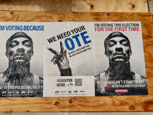 On a boarded up section of a brick wall, three posters. The outer two show Snoop Dogg's face, and the middle one shows a hand making a "V" gesture. Text of the 3 posters: 1. "I'm voting because I want to end police brutality." 2. "We need your vote. Black lives matter. Black votes matter. Register here. [QR code.]" 3. "I'm voting this election for the first time because I can't stand to see that punk in office another year." 