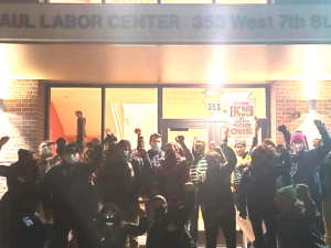 A group of people stands outside the St. Paul Labor Center, fists raised. It is dark out, and they are backlit.