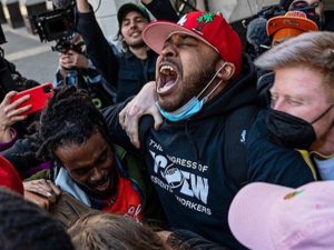 Tight crowd of people outdoors embracing and yelling in celebration. Most visible person in center, face upturned, wears a red baseball cap and a T-shirt that says The Congress of Essential Workers. Behind, many people hold up cameras capturing the moment.