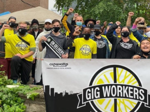 Young people of various races, fists in air, all masked. Some wear yellow "Gig Worker United" shirts and those in front hold a big banner with the group's logo and the initials of the Canadian postal union.