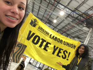 Inside a warehouse three young people smile as they unveil a bright yellow banner that says "Amazon Labor Union, Vote Yes"