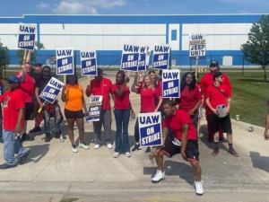 A group of Black and white men and women, most in red T-shirts, stand in front of the plant gates. Many carry blue and white "UAW on strike" picket signs.  