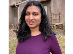 Sindhu poses outside in front of a barn. She has long black hair and is wearing a red shirt and a slight smile.