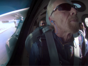 Screenshot of Richard Branson, wearing sunglasses and earphones, in a rocket. Two other crew members are visible behind him. On the left is a window through which you can see part of the outside of the rocket, labeled VIRGIN, and blue sky.