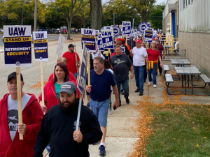 A dense line of picketers (at least 25 people are visible but it looks like there are more behind them) marches towards the camera on a leafy sidewalk alongside a factory. Many carry printed signs like "UAW on strike" and "Stand up for retirement security." Most visible faces are white but a few Black faces are also visible; there's a mix of women and men.