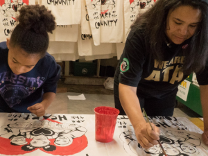 A child and an adult paint signs. Behind them are many more signs hanging already painted.