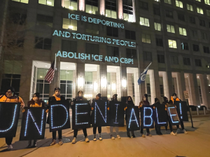 People holding lit signs spelling out "UNDENIABLE" stand in front of a building. On the building are projected the words: "ICE IS DEPORTING AND TORTURING PEOPLE! ABOLISH ICE AND CBP!"