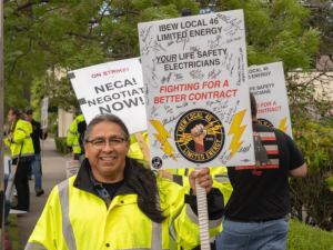 Picketers picket on a sidewalk. Person in foreground, wearing a safety-yellow jacket, smiles at the camera. This person has brown skin, glasses, hair in a ponytail, and holds a Local 46 picket sign printed with a limited energy of a fist holding bolts of lightning and the words "Your Life Safety Electricians, Fighting for a Better Contract." The sign appears to be autographed by many other strikers. Another printed sign visible in background says "On strike! NECA! Negotiate now!"