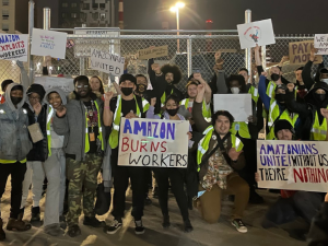 Crowd of excited owrkers stands outside warehouse in the dark holding up handmade signs. Most prominent ones read "Amazon burns workers," "Amazon exploits workers," "Amazonians unite! Without us they're nothing."