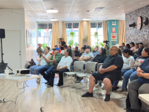 In a bright sunny room, a man in a black polo shirt and jeans stands on the left, speaking into mic, while about 30 people seated in chairs on the right listen. Most or all appear Latino/a. 