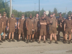 About 20 UPS workers in brown shorts uniforms pose for the camera together in a sunny parking lot. Many are smiling.