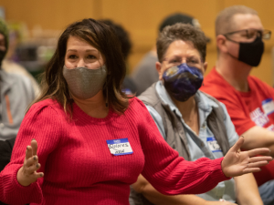 People in masks and nametags seated inside. One woman in red sweater is gesturing while speaking; others are listening.