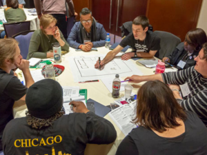 People sit around a big table, leaning in. One is drawing a workplace map on big paper, while another points something out. They're in a hotel conference room. Other table groups are visible behind them.