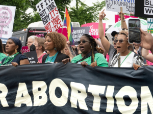 Women march behind a giant banner. The part we can see reads "OUR ABORTION." Other signs visible say "If it's not your body, it's not your choice" and "Our bodies, our future."