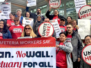 Workers in red UNITE HERE shirts and carrying round matching signs stand on steps outside. Those in front hold a large printed banner: "Tech Cafeteria Workers Say... No Ban, No Wall, Sanctuary for All."