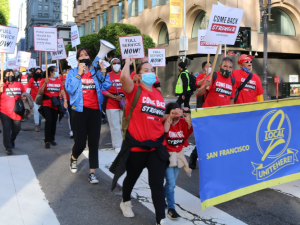 Workers march in downtown streets behind a big blue banner that says "UNITE HERE Local 2." They are wearing red T-shirts that say "Come back stronger" and some carry picket signs saying "Full service now!" or "Hotel rooms should be cleaned every day."  Most wear masks, one holds a bullhorn, and there is a child near the front of the crowd.