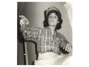 Black-and-white photo looks upward at a woman in a plaid shirt and bedazzled hardhat, gesturing and speaking