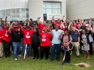 A large group of Black and white women and men stands on grass outside the Mercedes-Benz Training Center, smiling, many with fists in the air. Many wear red T-shirts with a UAW logo and the words "Mercedes Workers United."