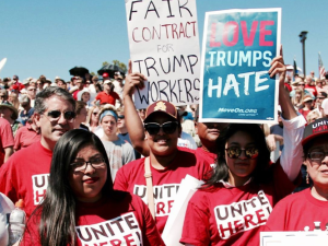 Workers in red UNITE HERE shirts pose at a huge rally. One handlettered sign says "Fair contract for Trump workers" and another printed sign says "Love Trumps Hate."