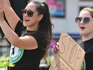 Two young women in Starbucks Workers United T-shirts stand outdoors, holding up cardboard signs, mouths open to yell or chant.