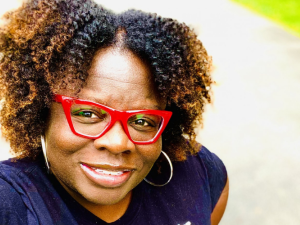 Close-up selfie of Petal Robertson, a Black woman with red glasses, smiling into the camera, outdoors